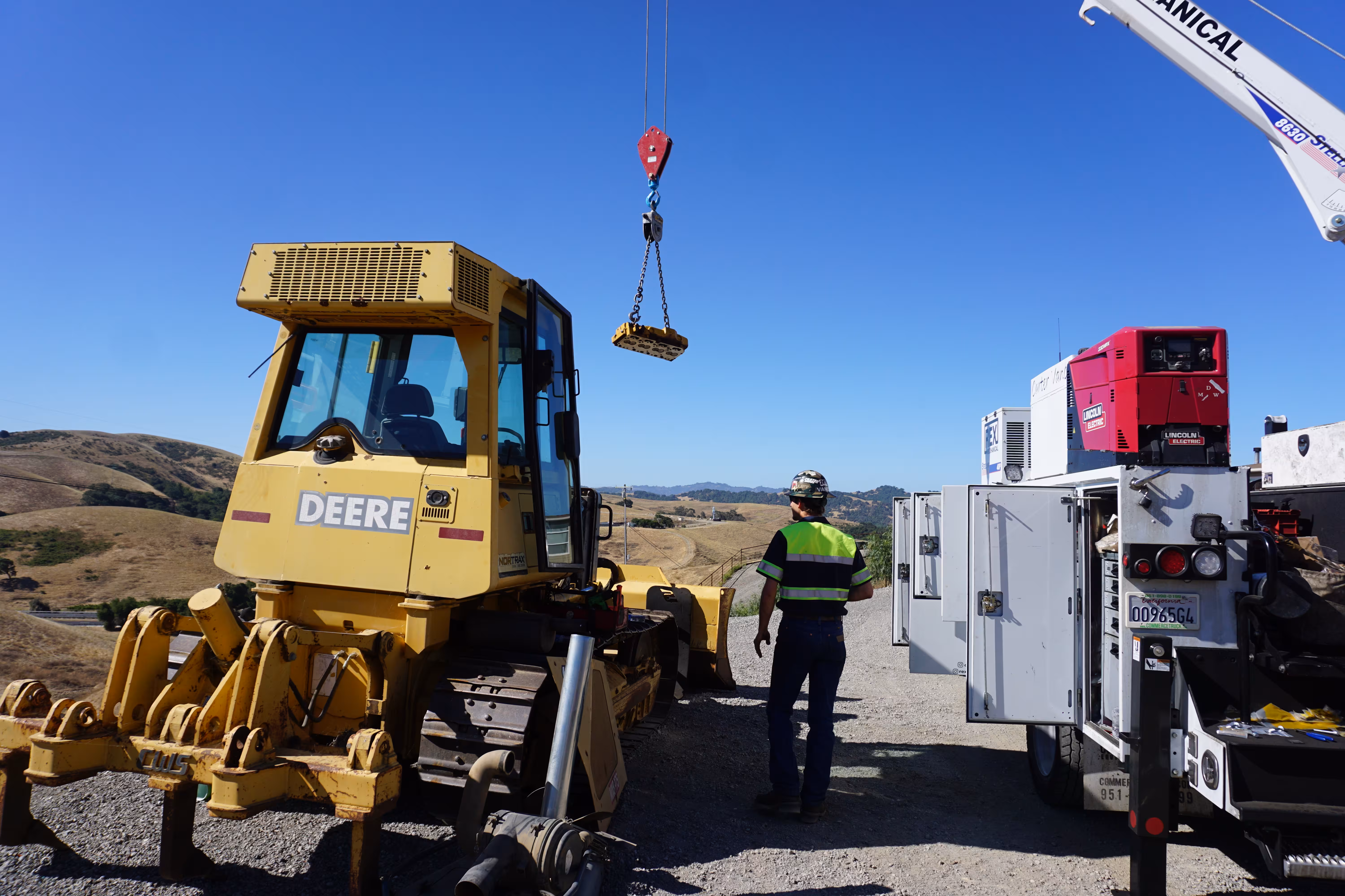 A service truck's boom crane lifts a component beside a John Deere dozer on a hilltop job site