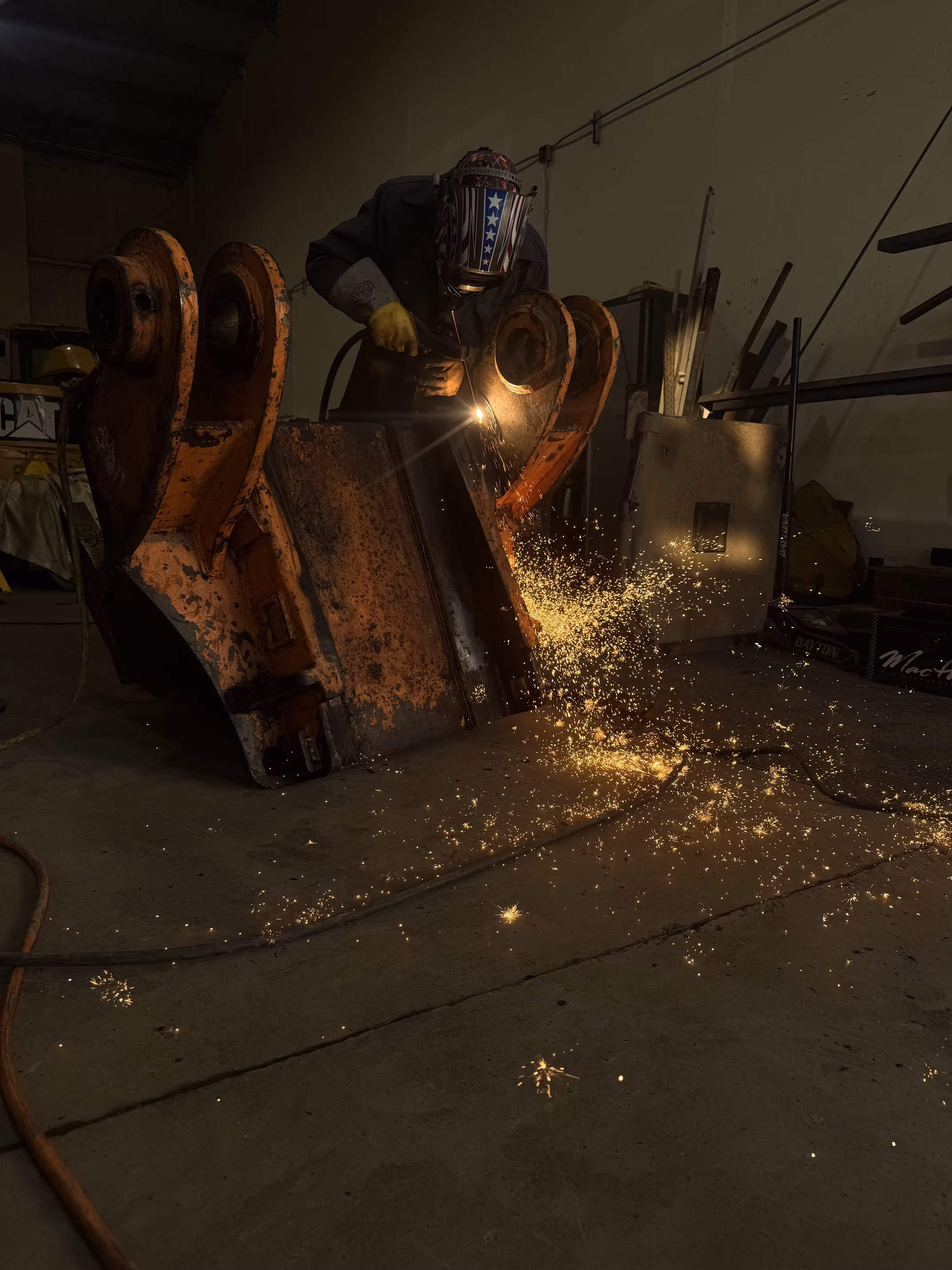 A technician welds a pin boss on a large excavator bucket, sparks arcing across the shop floor