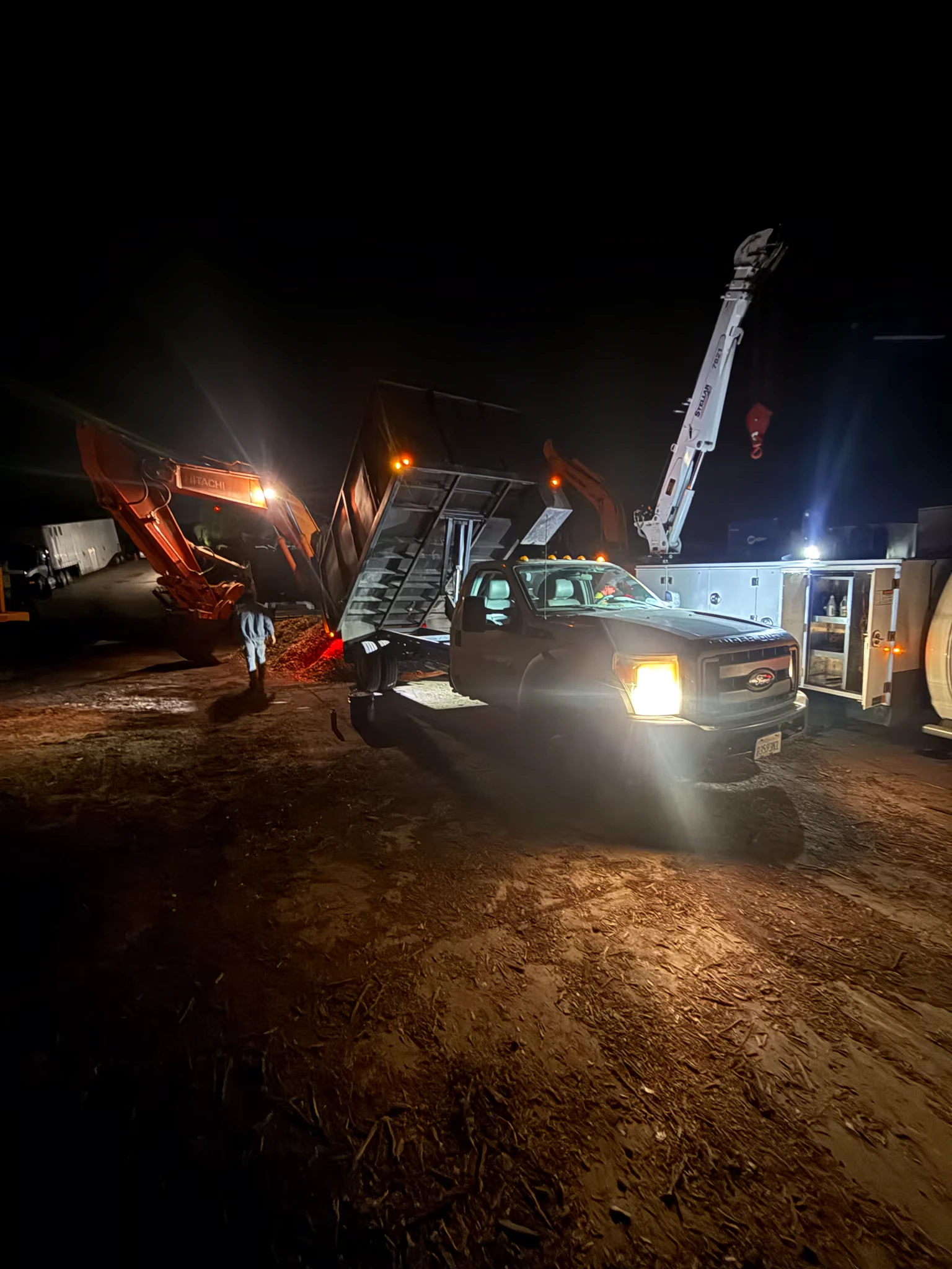 A service truck with a boom crane parked at night beside a raised dump bed and a Hitachi excavator