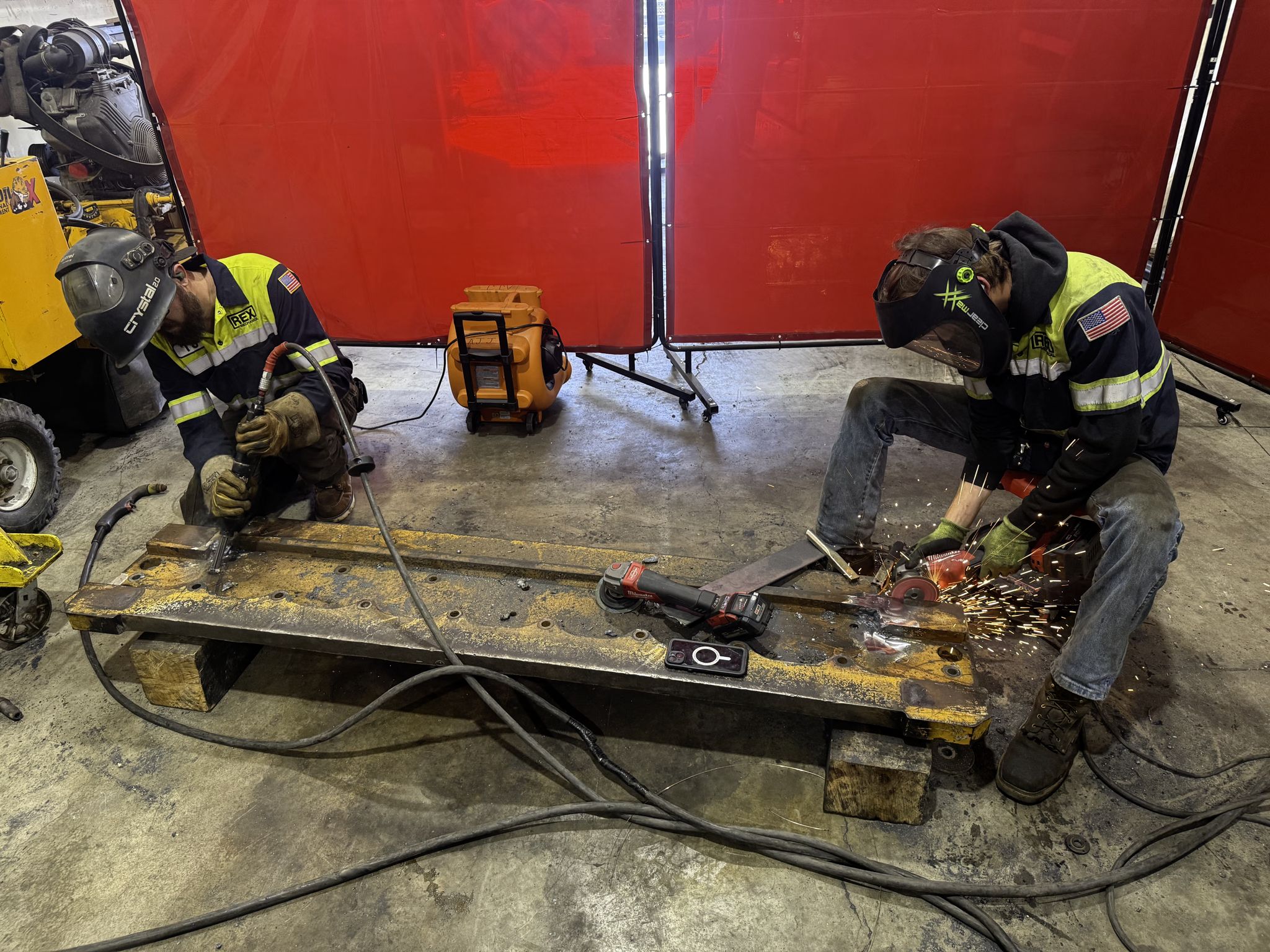 Two technicians weld and grind a steel beam on blocks in the shop, one in a REX Mechanical jacket