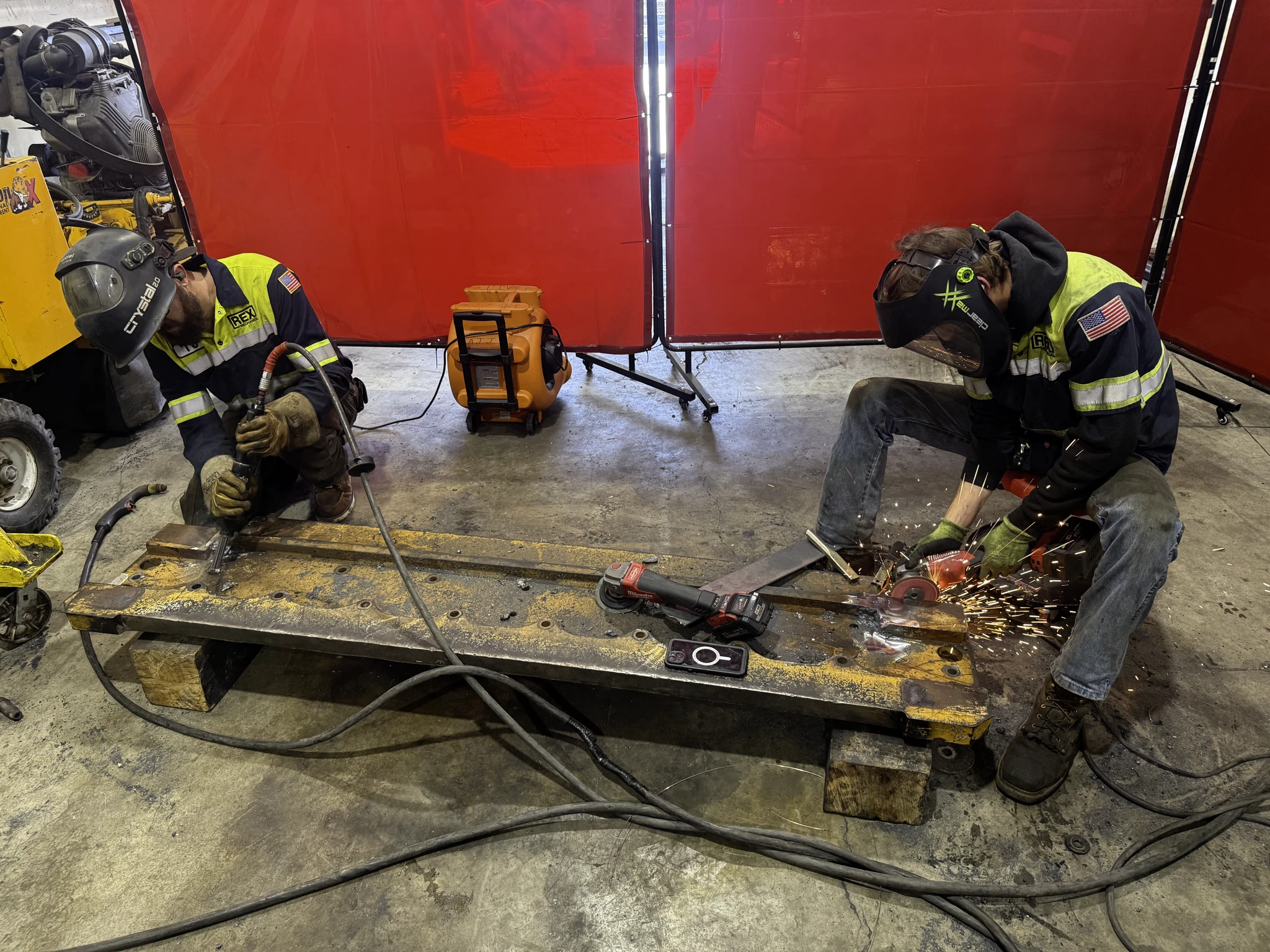 Two technicians weld and grind a steel beam on blocks in the shop, one in a REX Mechanical jacket