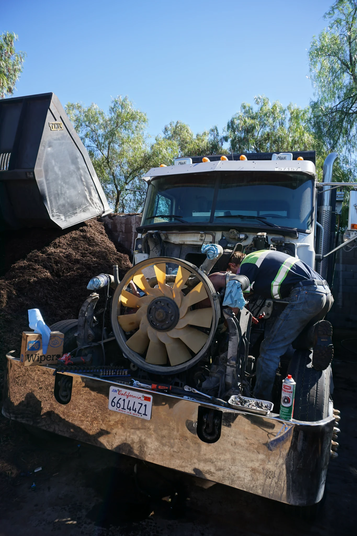 A technician leans into the engine bay of a Mack dump truck past the exposed cooling fan