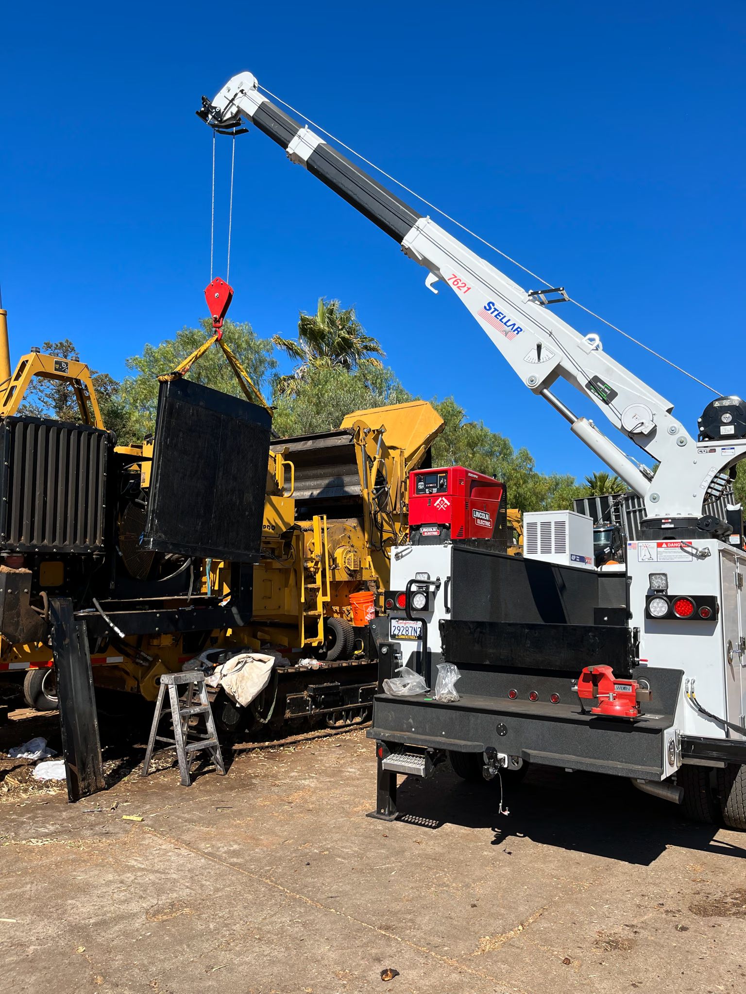 A truck-mounted Stellar crane lifts a steel panel off a yellow tracked grinder in a work yard