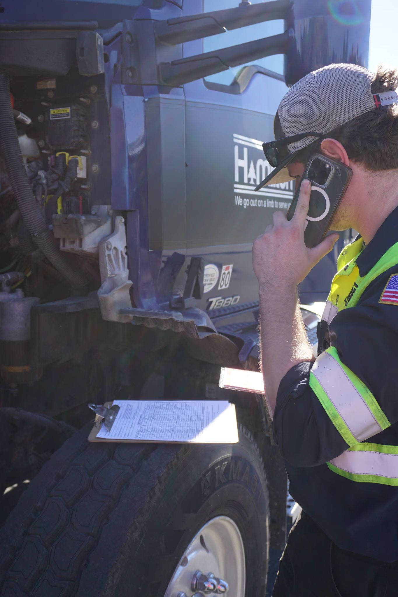 A technician takes a call at the open engine bay of a Kenworth T880, inspection sheet on a clipboard