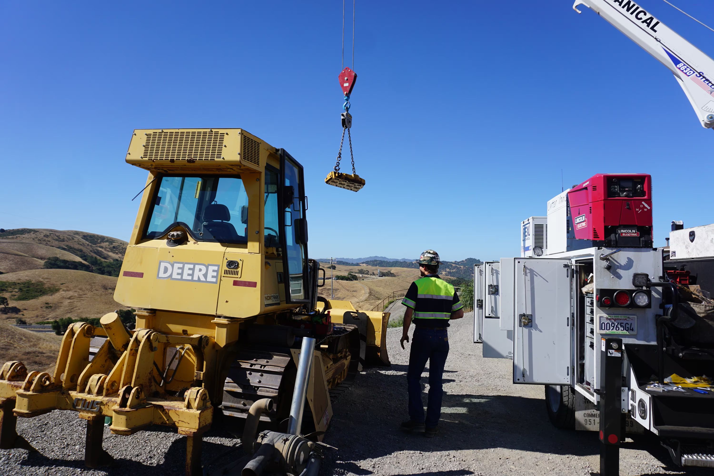 A service truck's boom crane lifts a component beside a John Deere dozer on a hilltop job site