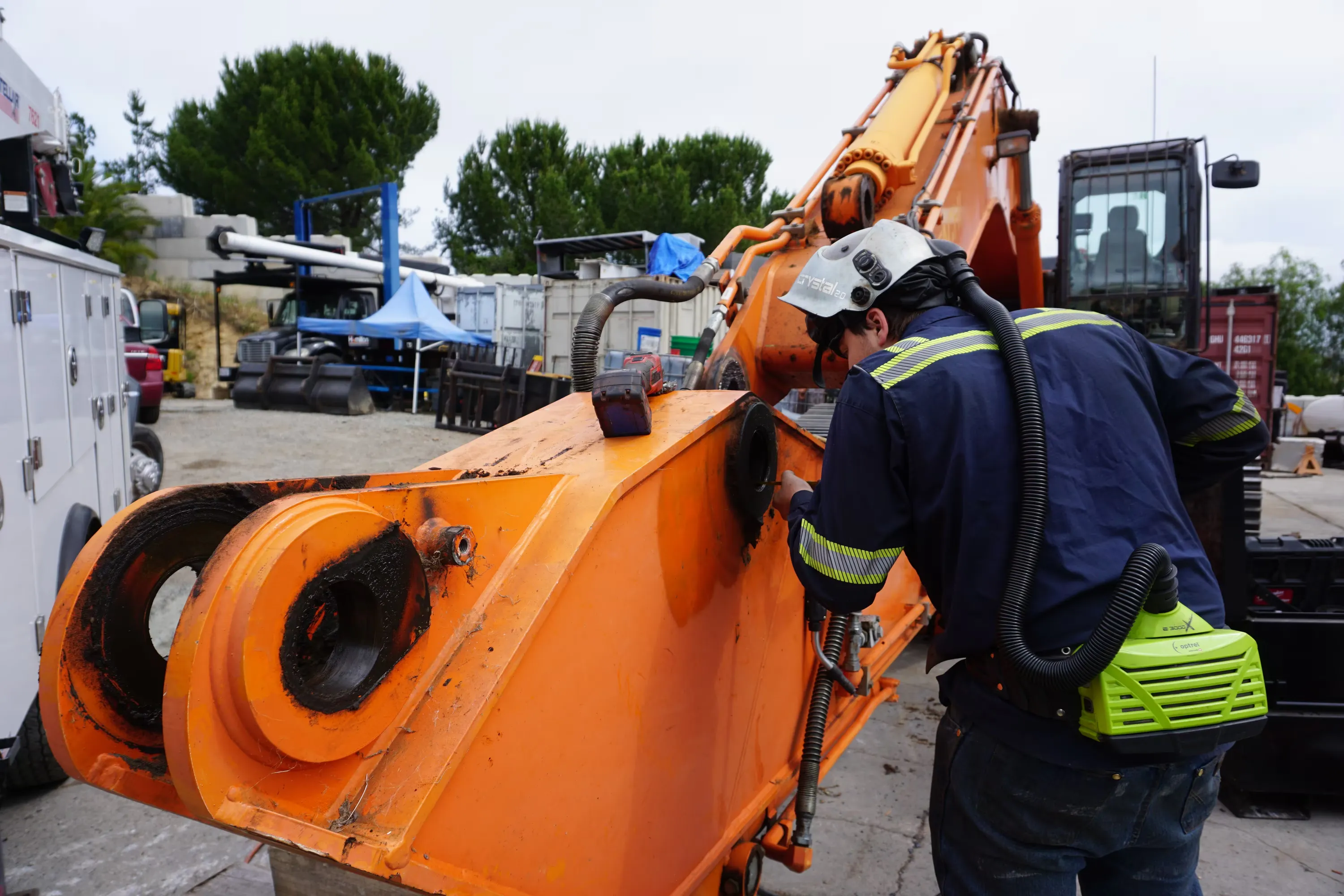A technician in a respirator hood works on the worn pin bores of an orange excavator boom