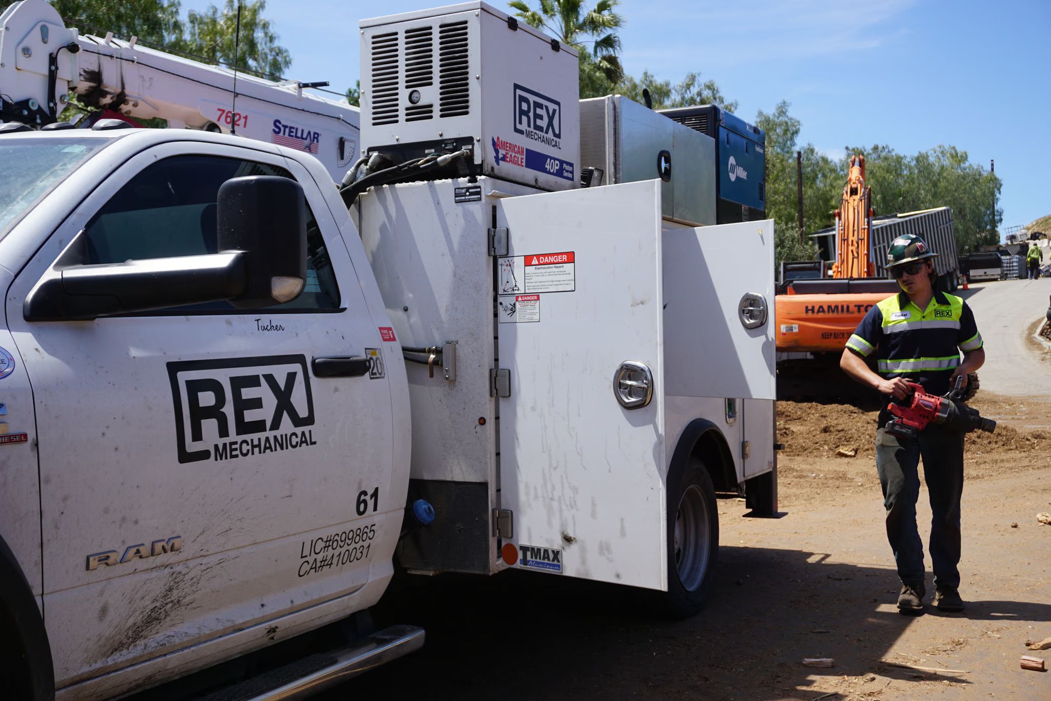 A technician carries an impact wrench past a REX Mechanical service truck at a dirt job site