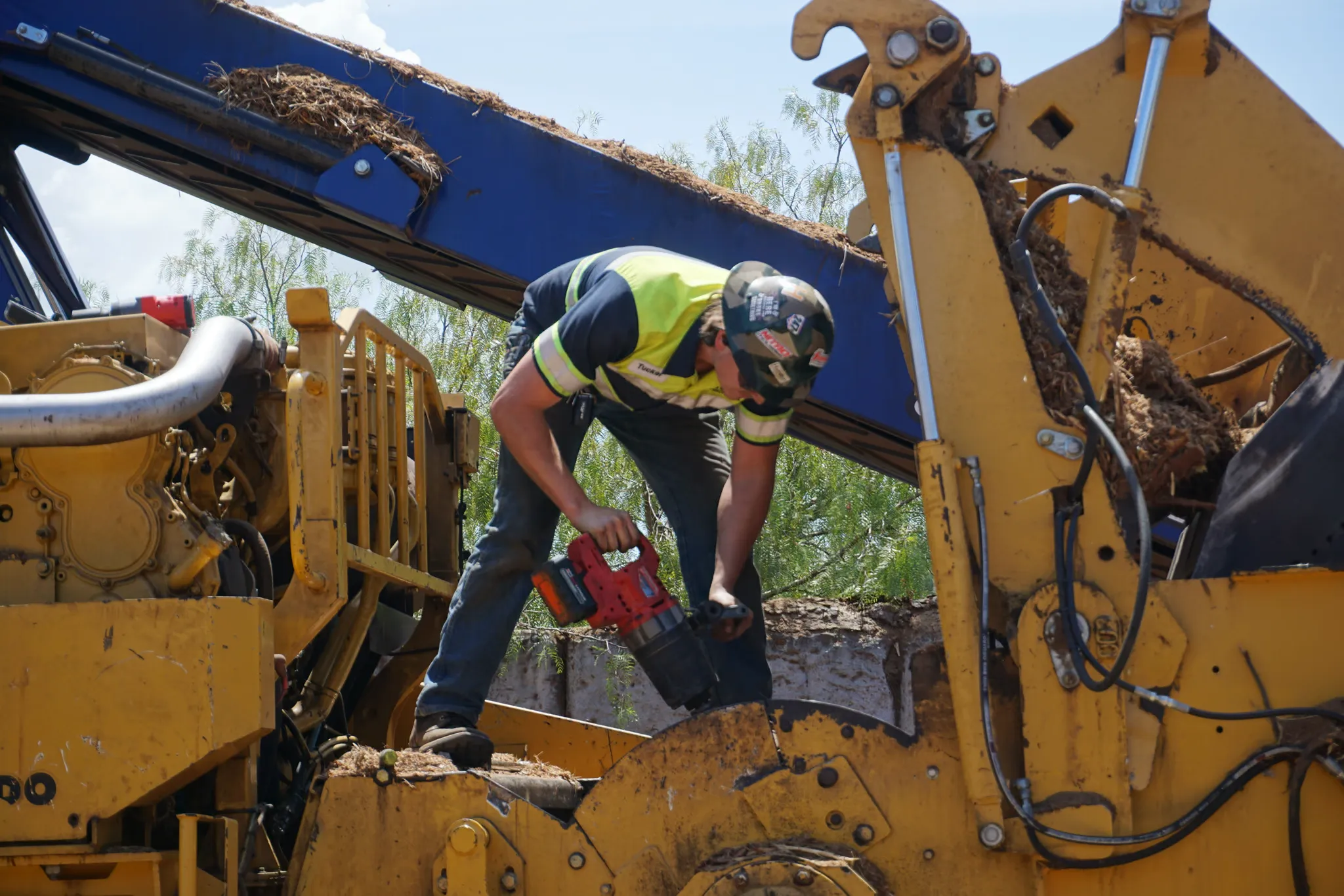 Technician bent over a yellow wood grinder, greasing it with a cordless grease gun