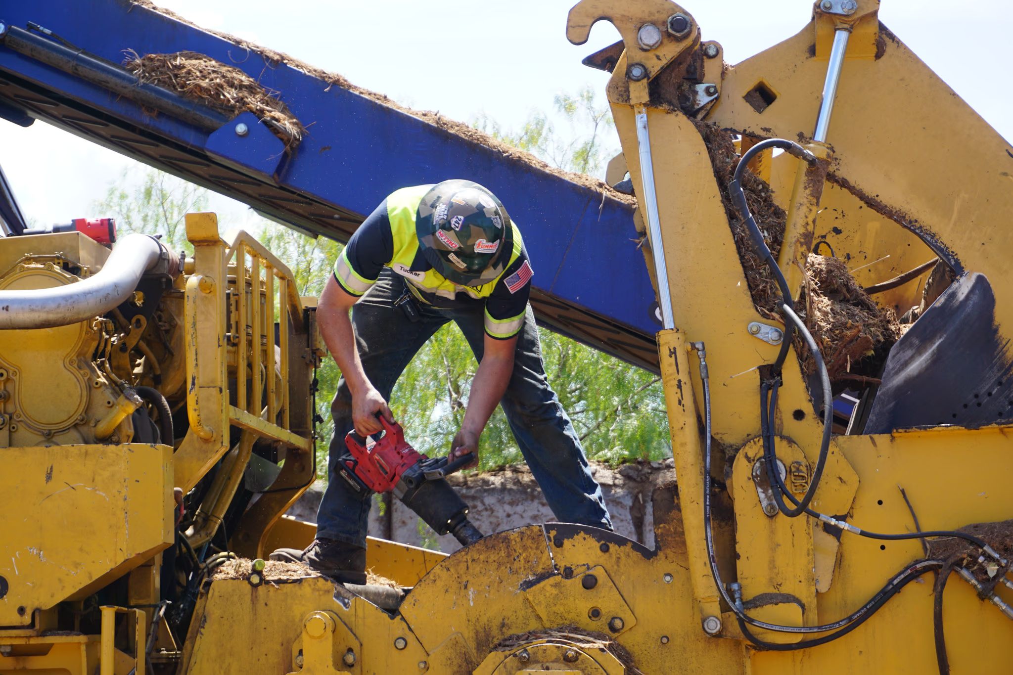 Technician with a cordless grease gun servicing a yellow wood grinder on a jobsite