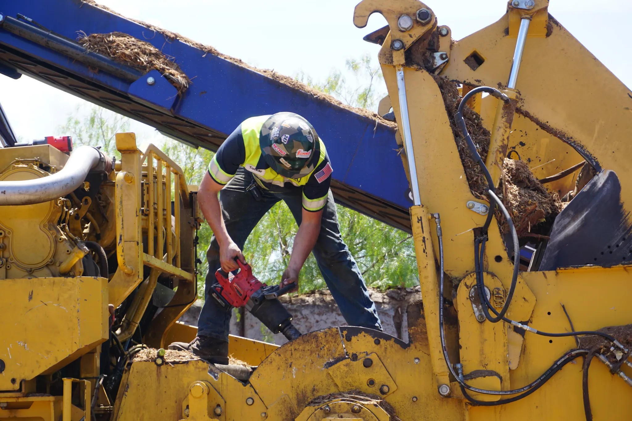 Technician with a cordless grease gun servicing a yellow wood grinder on a jobsite