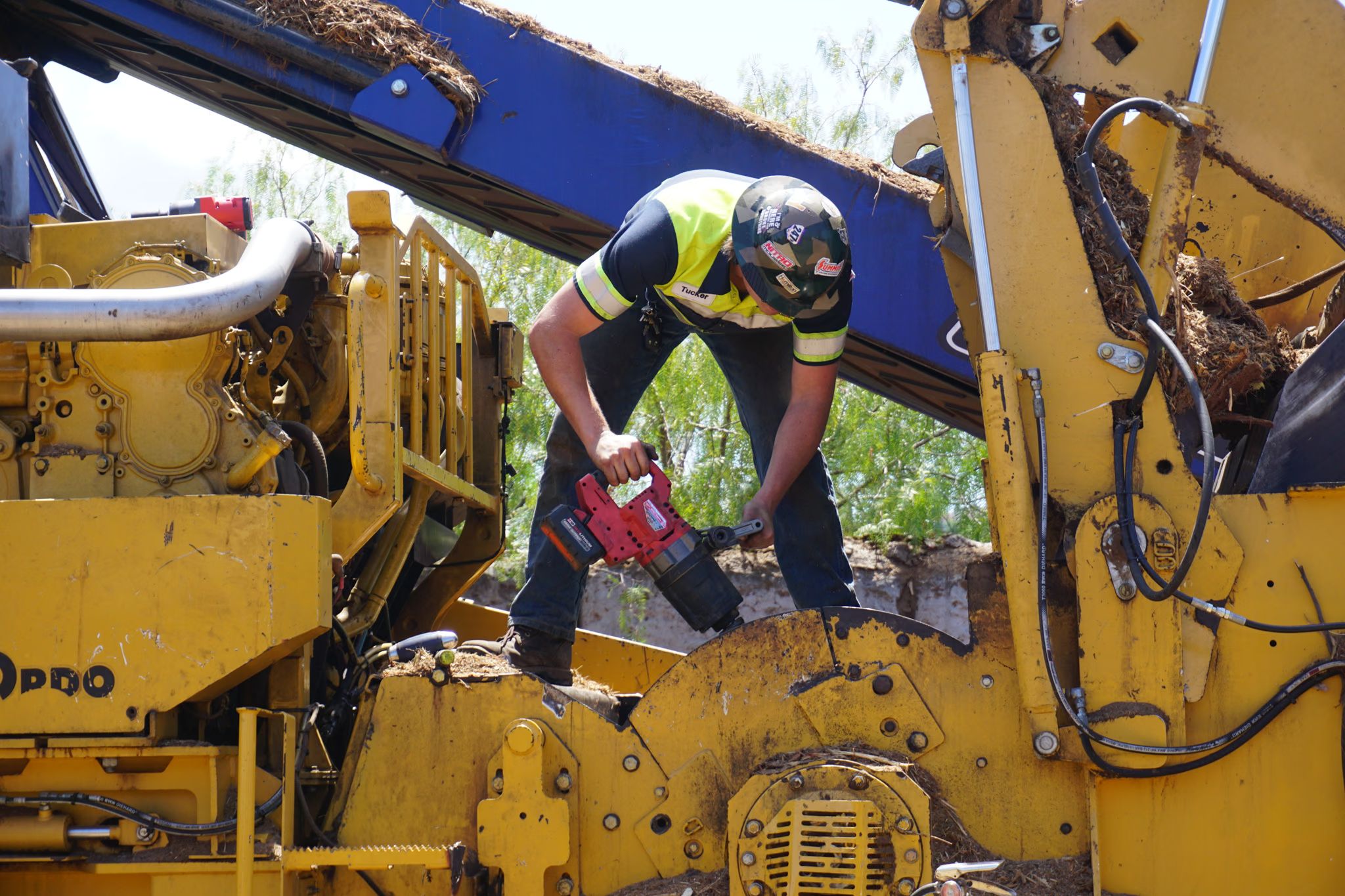 Technician stands over a yellow wood grinder, cordless grease gun and hose in hand