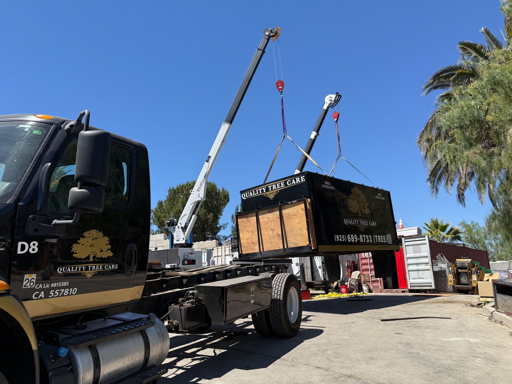 Two truck-mounted booms hold a chip box in the air above an empty flatbed chassis in a yard