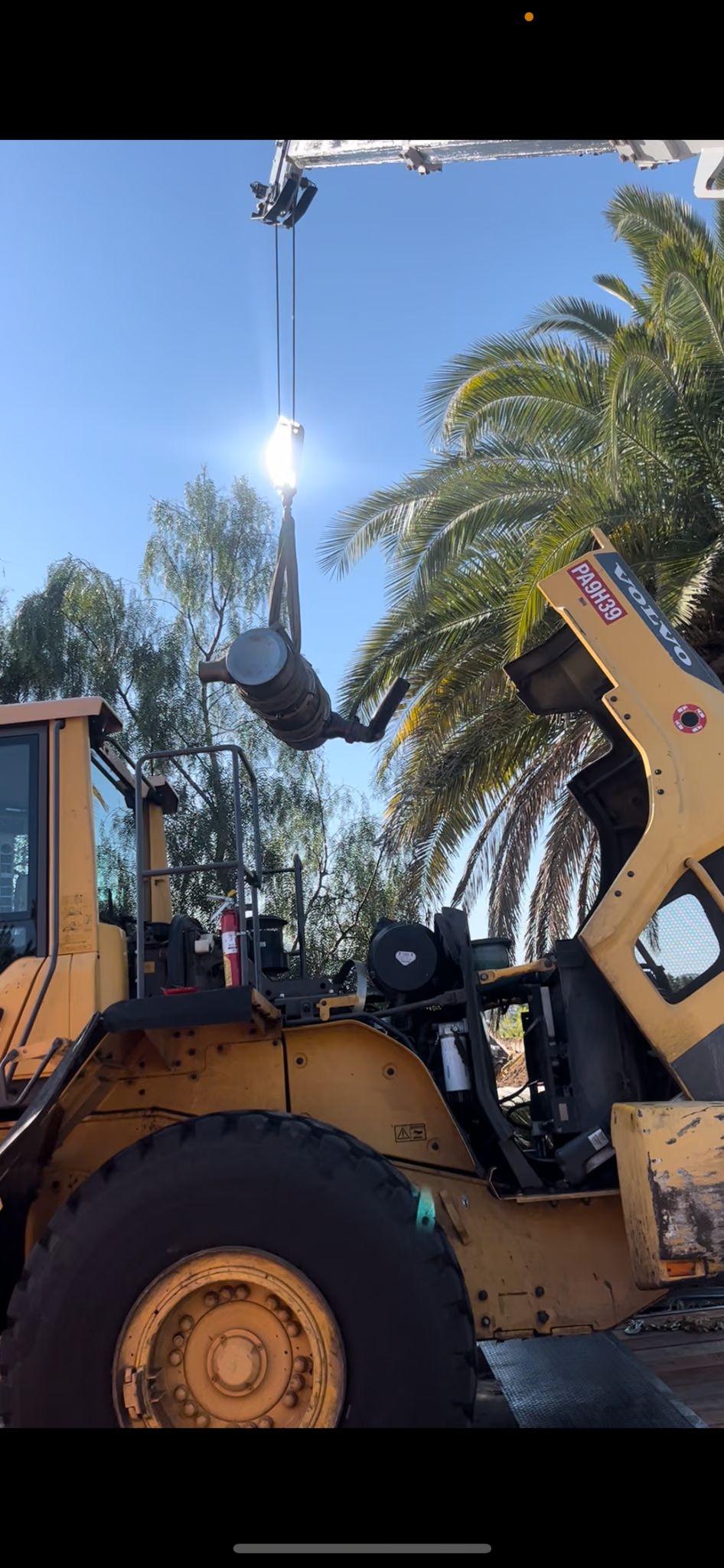 A crane sling lifts a large cylindrical component from the open engine bay of a Volvo wheel loader