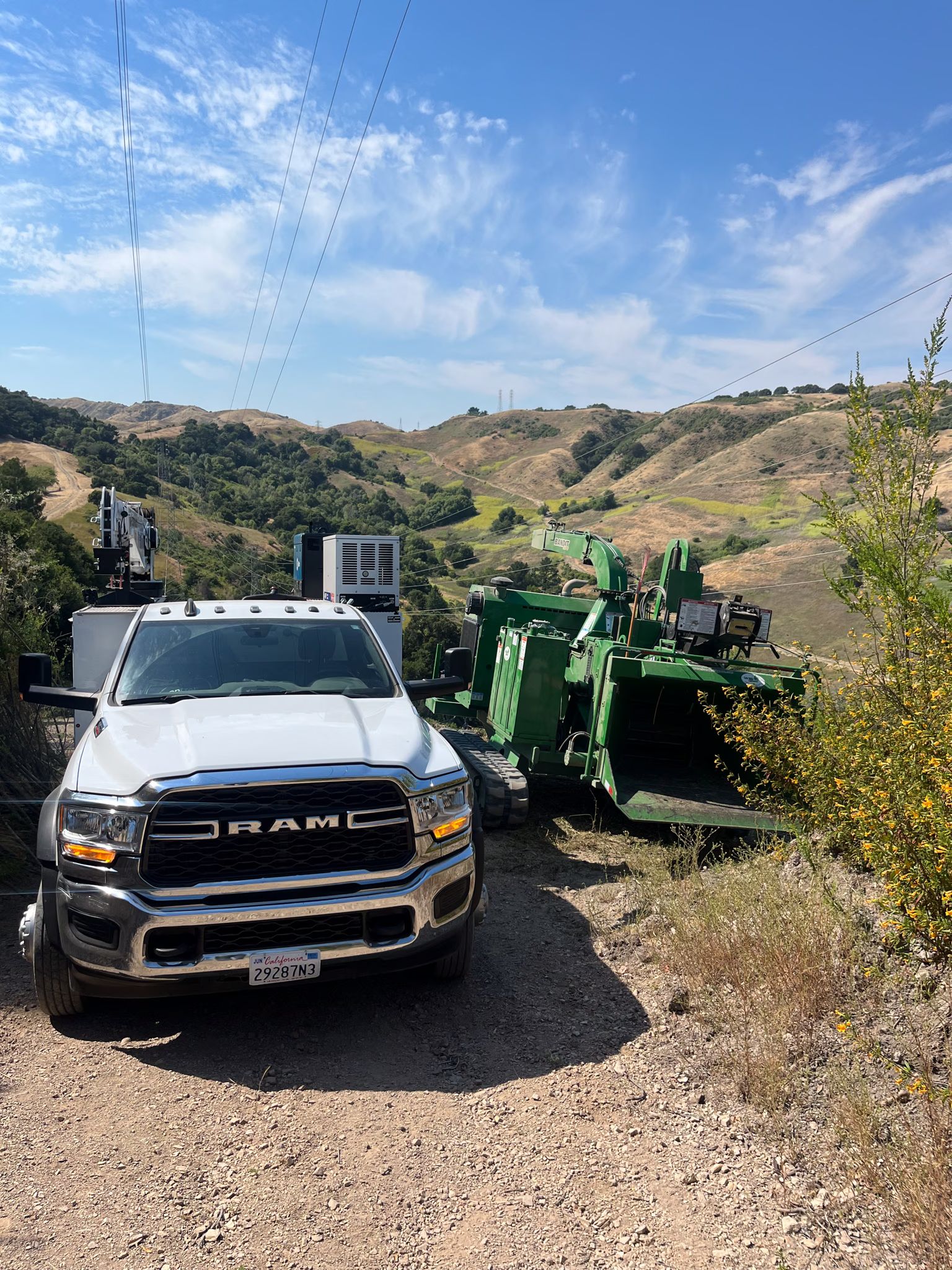 Ram service truck with a bed crane on a dirt ridge road beside a large green wood chipper