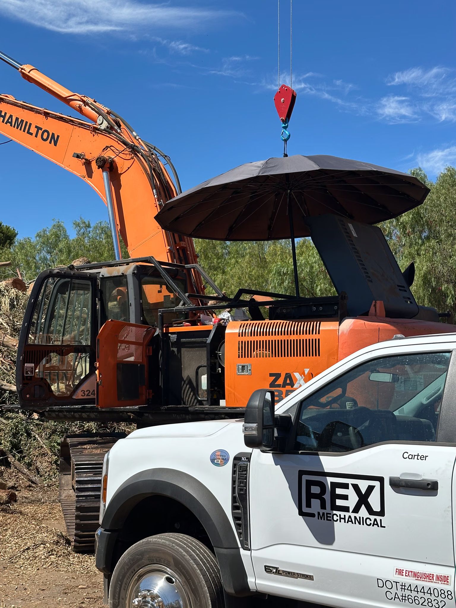 REX Mechanical truck at an excavator with its engine bay open, shaded by an umbrella on a crane hook